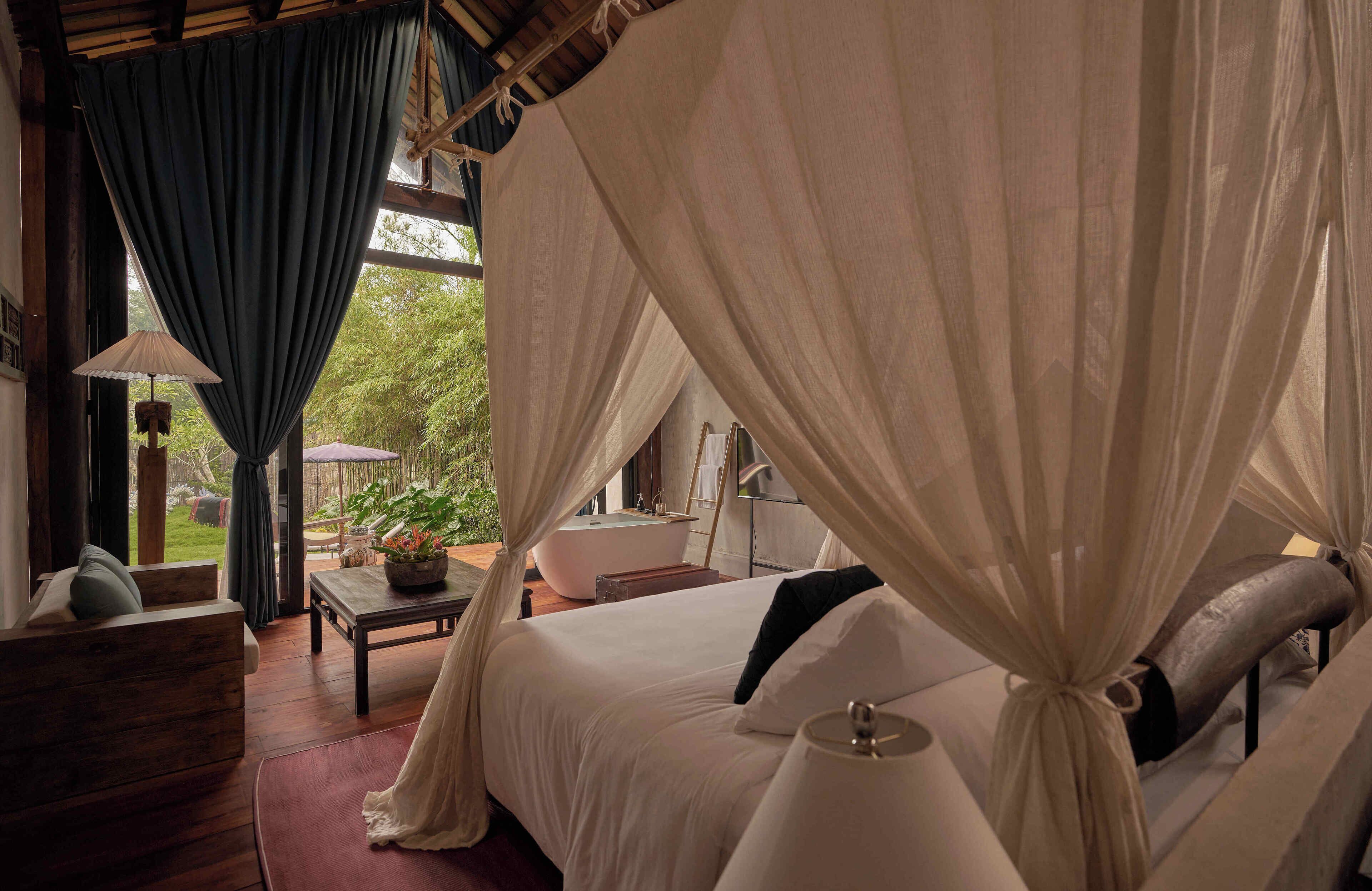 A bedroom with a canopy bed, a wooden nightstand, a table with a lamp, and a view of the outdoors.