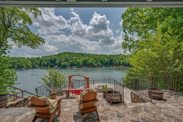 A patio with Adirondack chairs, a wooden table, and a stone floor overlooking a lake surrounded by trees. - M & M Rockhouse | Pet-Friendly Lake Cottage on Lake Lure with Private Lake Access and Fire Pit (Lake Lure)