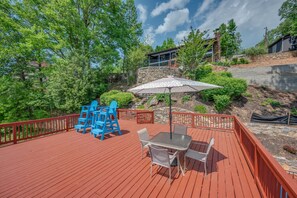 A wooden deck with outdoor furniture, a stone wall, and a view of a house in the background. - M & M Rockhouse | Pet-Friendly Lake Cottage on Lake Lure with Private Lake Access and Fire Pit (Lake Lure)