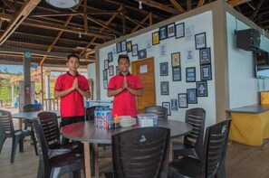 Two individuals in red shirts standing in a covered dining area with tables and chairs, framed pictures on the walls, and a speaker mounted on the wall. - SBS Resort by Zuzu (Galang)