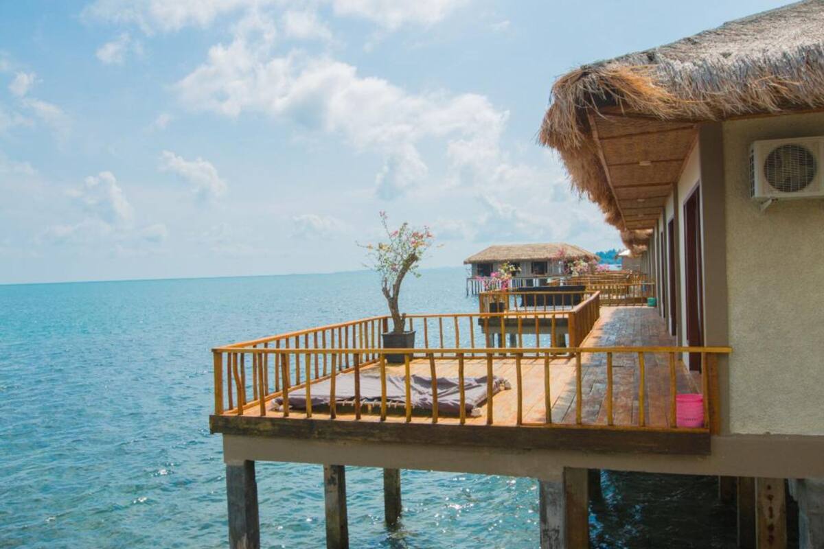 A seaside overwater bungalow with a thatched roof, a balcony with a railing, and a view of the ocean.