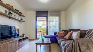 Ein Wohnzimmer mit Fernseher, Holzregalen, einer Couch, einem Couchtisch und einer Schiebetür zu einer Terrasse.