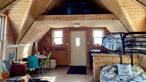 A cozy attic bedroom with a sloped wooden ceiling, a bunk bed, a desk, and a dining area with chairs and a table. - Off grid solar a-frame cabin Northwoods Farm Stay Hundreds of Acres to Explore (Prentice)