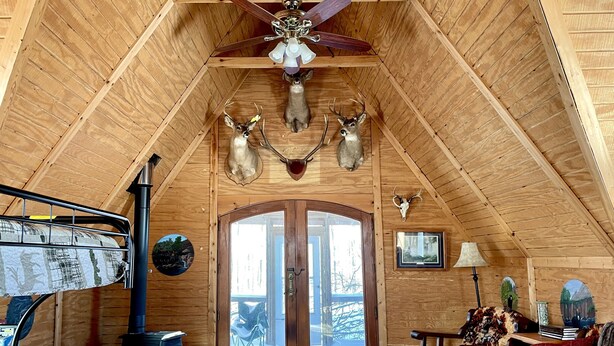 A log cabin interior with a high ceiling, wooden walls, a fireplace, a large window, and deer antlers mounted on the wall. - Off grid solar a-frame cabin Northwoods Farm Stay Hundreds of Acres to Explore (Prentice)