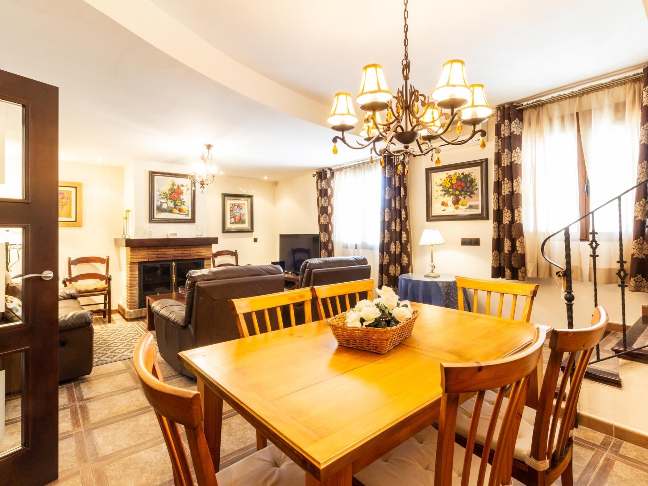 A dining area with a wooden table and chairs, a chandelier, a fireplace, and a television in a living room with a sofa and a chair.
