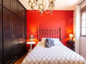 Ein Schlafzimmer mit roter Wand, einem Bett mit gemustertem Überwurf, einer Chandel üster und einem Nachttisch mit Lampe.