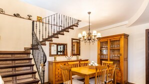 Un comedor con una mesa y sillas de madera, una araña, un aparador con cristalería y una escalera con barandilla negra.