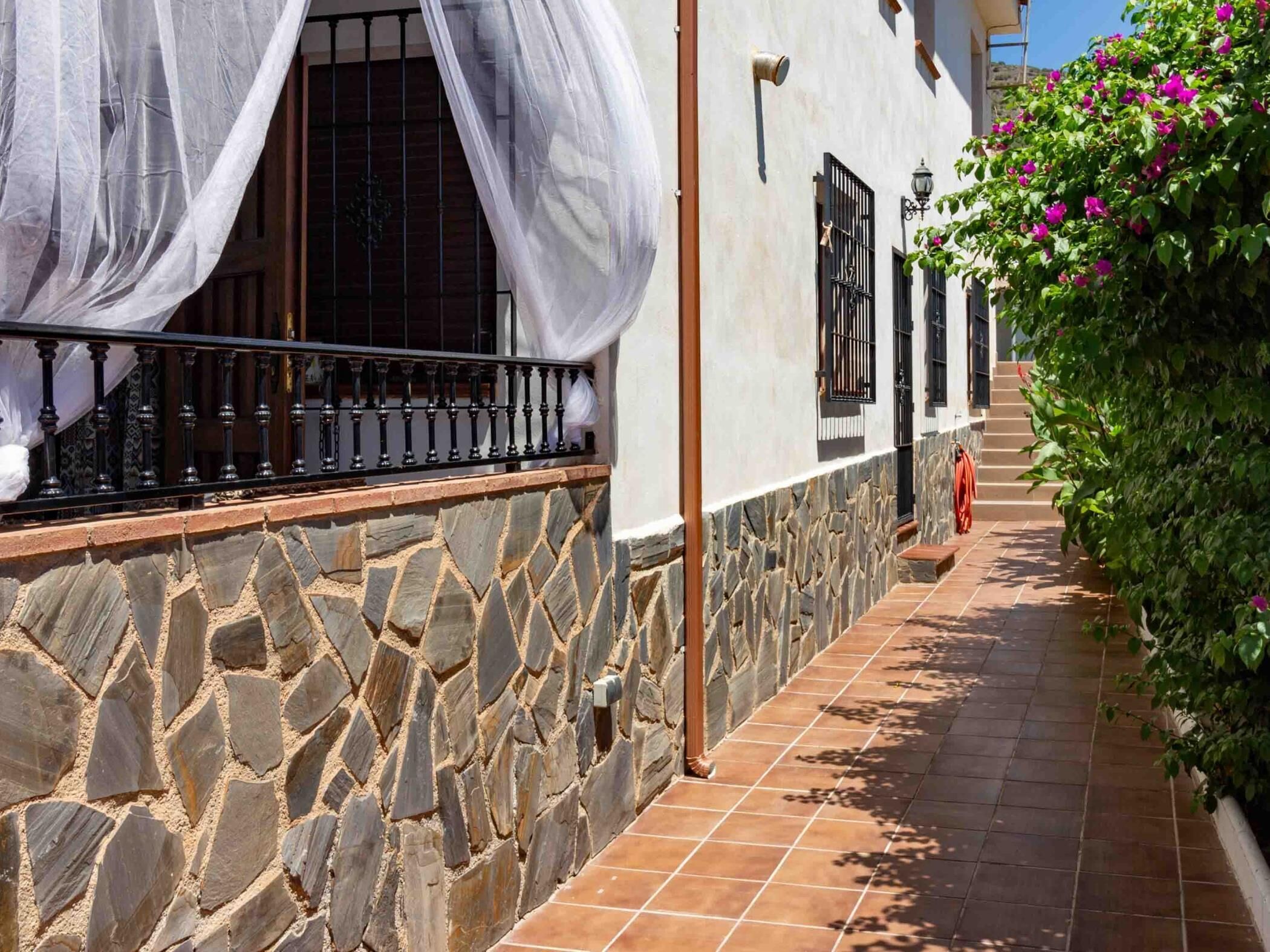 Un edificio de paredes de piedra con un sendero de baldosas, un balcón con barandilla negra y una ventana con cortina blanca.