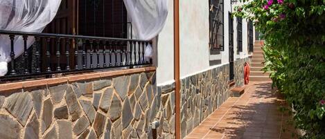 Un edificio de paredes de piedra con un sendero de baldosas, un balcón con barandilla negra y una ventana con cortina blanca.