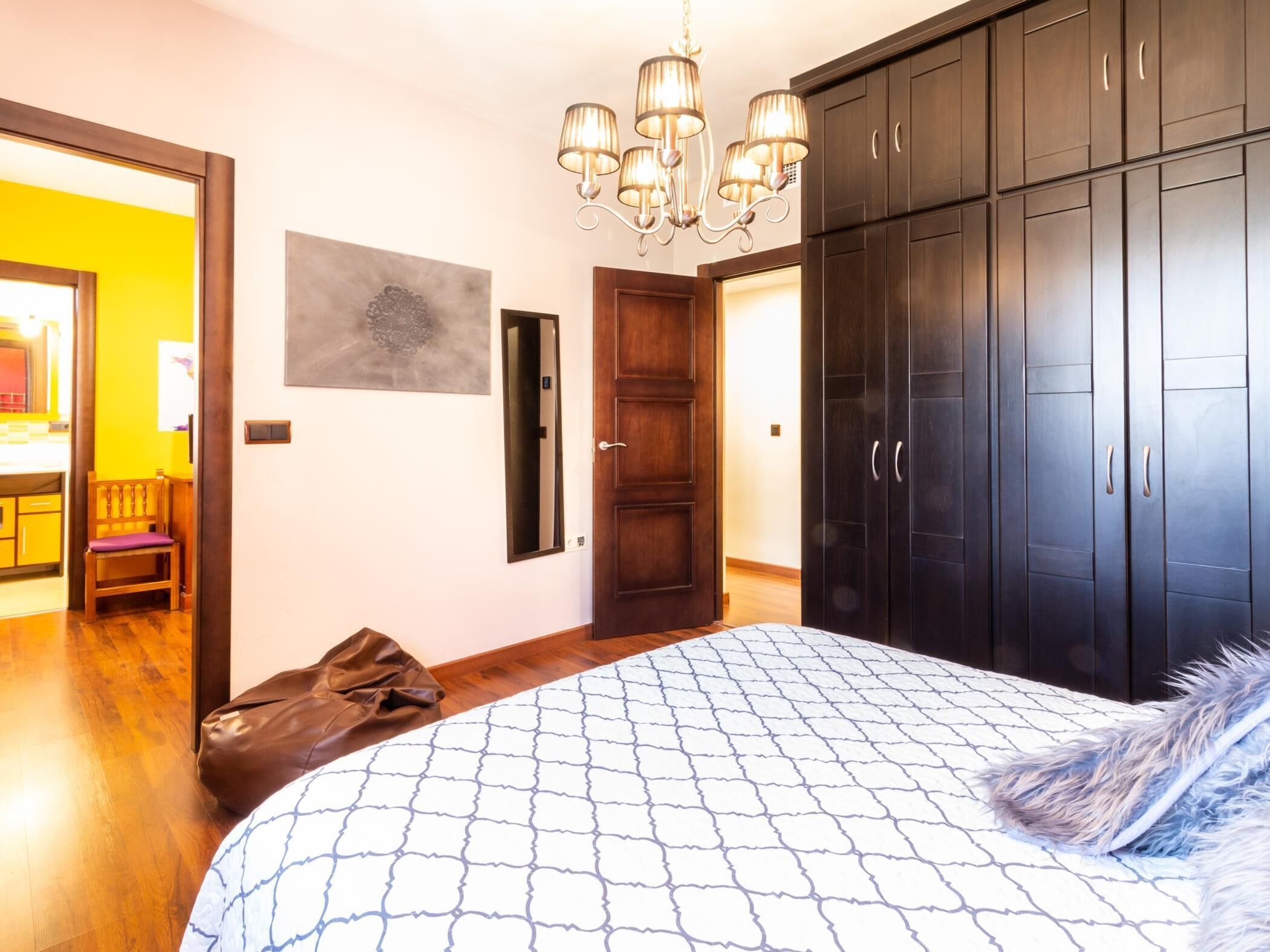 A bedroom with a large bed, a wooden chair, a brown bag, a chandelier, and dark wooden wardrobes.