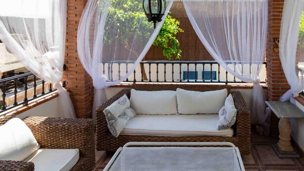 Un espacio al aire libre techado con un sofá y sillas de mimbre, un farol colgante y vistas a la vegetación a través de cortinas translúcidas.