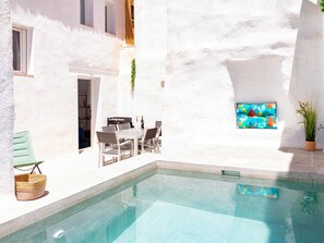 Ein Poolbereich mit einer weißen Wand, einem kleinen Tisch mit Stühlen und einem Fenster mit einem farbenfrohen Kissen.