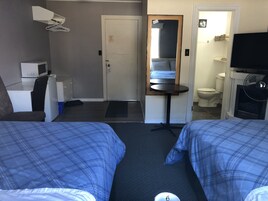 A hotel room with two beds, a desk, a chair, a microwave, a small refrigerator, a mirror, a fan, and a toilet.