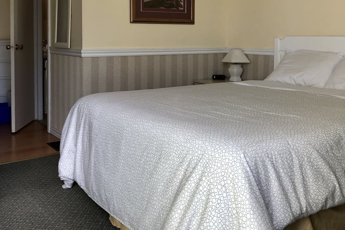 A hotel room with a large bed, a painting on the wall, a bedside table with a lamp, and a door leading to a bathroom.