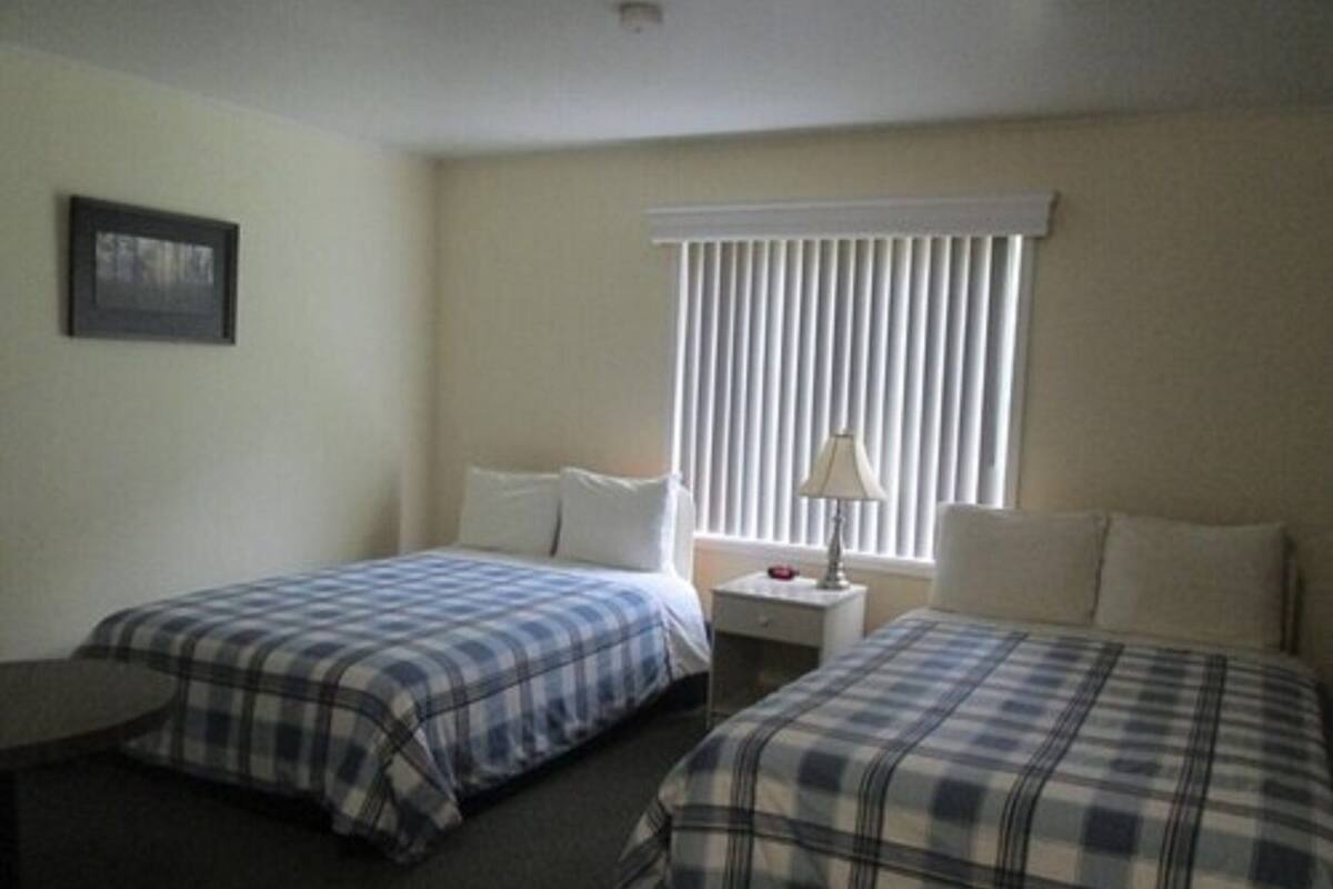 A hotel room with two beds, a nightstand with a lamp, a small table, and a window with blinds.