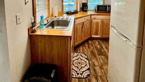 A compact kitchen with wooden cabinets, a stainless steel sink, a microwave, and a refrigerator. There are three windows with blinds, a ceiling light, and a patterned rug on the floor.