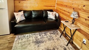 A cozy room with a wooden ceiling, a black leather sofa with floral cushions, a small table with a lamp, and a white refrigerator.