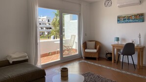 A modern living room with a sofa, a round table, a wicker chair, and a dining table with chairs. There is a balcony with a view of buildings and a palm tree. - Townhouse, surrounded by gardens and swimming pools (Cala d'Or)