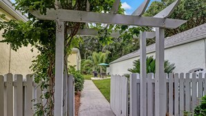 Un chemin menant à une maison entourée d’une clôture en bois peinte en blanc, agrémentée d’une pergola et bordée d’une pelouse.