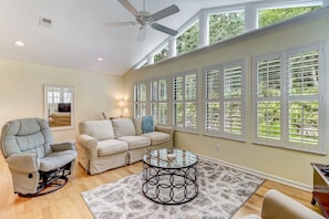 A living room with a recliner, a sofa, a glass coffee table, and large windows with shutters.