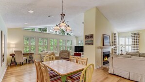 Un salon spacieux avec une cheminée, un coin repas avec une table et des chaises en bois, et de grandes fenêtres équipées de stores.