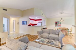 A spacious living room with a grey sofa set, a glass coffee table, and a kitchen area with red cabinets and a white refrigerator.