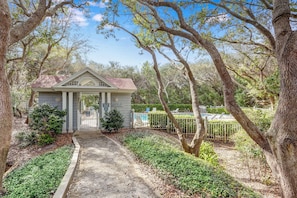 A small, single-story building with a gabled roof, surrounded by mature trees and a fenced pool area.