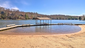 A lakeside beach with a wooden dock and benches, surrounded by trees and buildings.