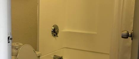 Un baño con inodoro, bañera y ducha. Hay toallas en un estante y un rollo de papel higiénico en la pared.