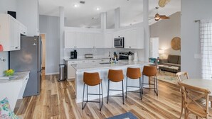 Une cuisine moderne avec des meubles blancs, un îlot central doté de tabourets, un ventilateur de plafond et un coin repas avec une table et des chaises.