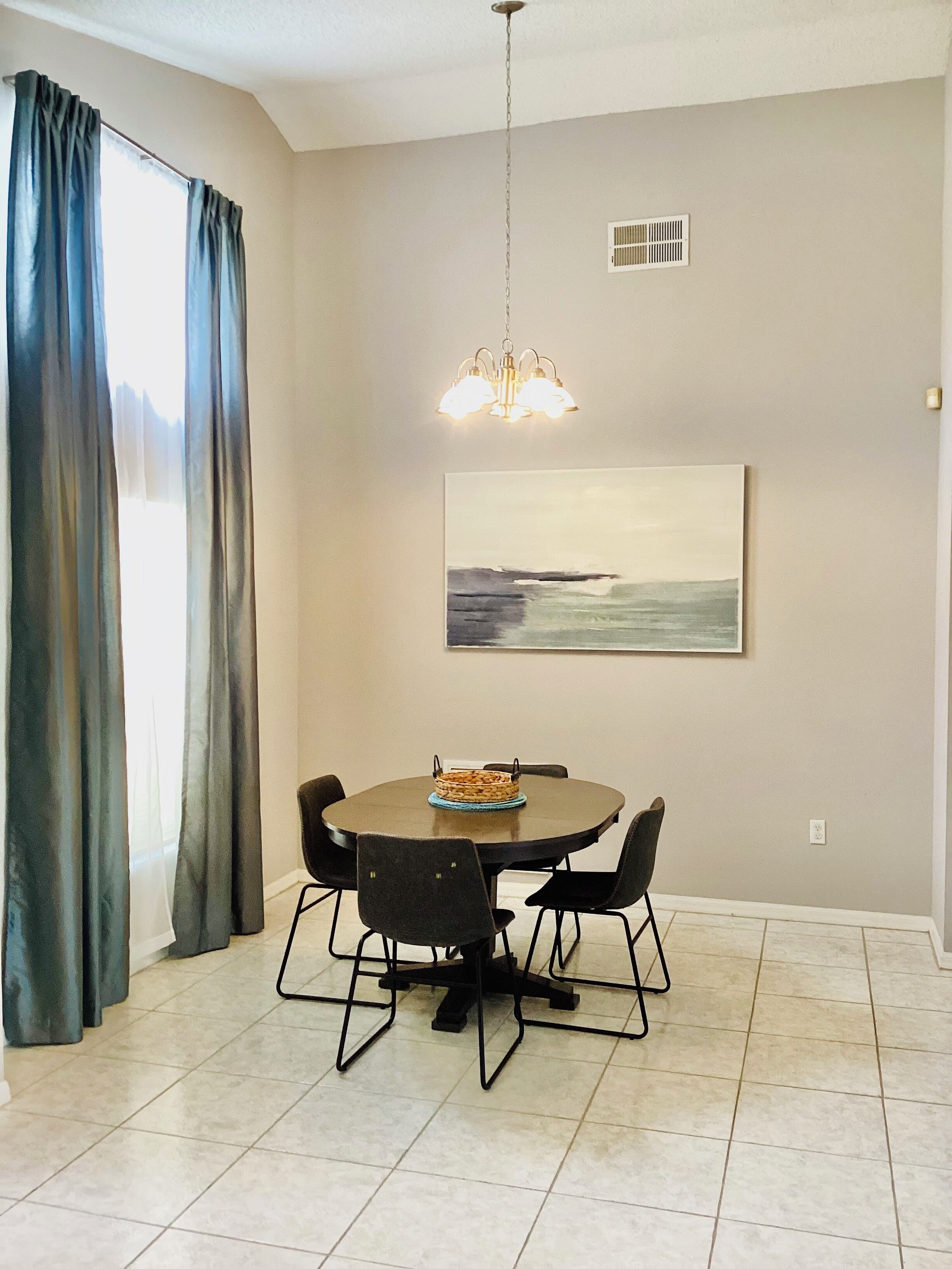 A dining area with a round table, four chairs, a chandelier, a painting on the wall, and a window with curtains.