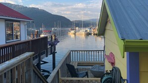 Een houten steiger die uitkomt bij een jachthaven met boten, een terras met stoelen, en een kleurrijk gebouw met een rood dak.