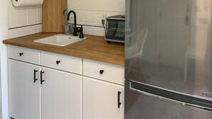 Een kleine keuken met witte kastjes, een houten aanrecht, een roestvrijstalen koelkast, een broodrooster en een plank met servies en glazen.