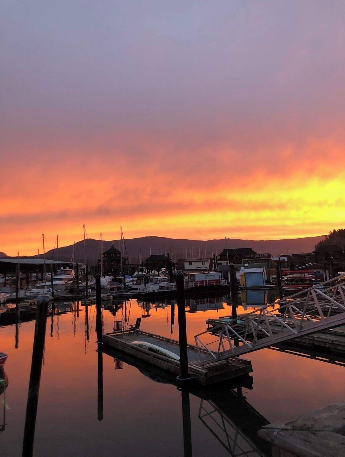 Un port de plaisance avec des bateaux amarrés, une jetée en bois et une surface d’eau calme qui reflète le ciel au coucher du soleil.