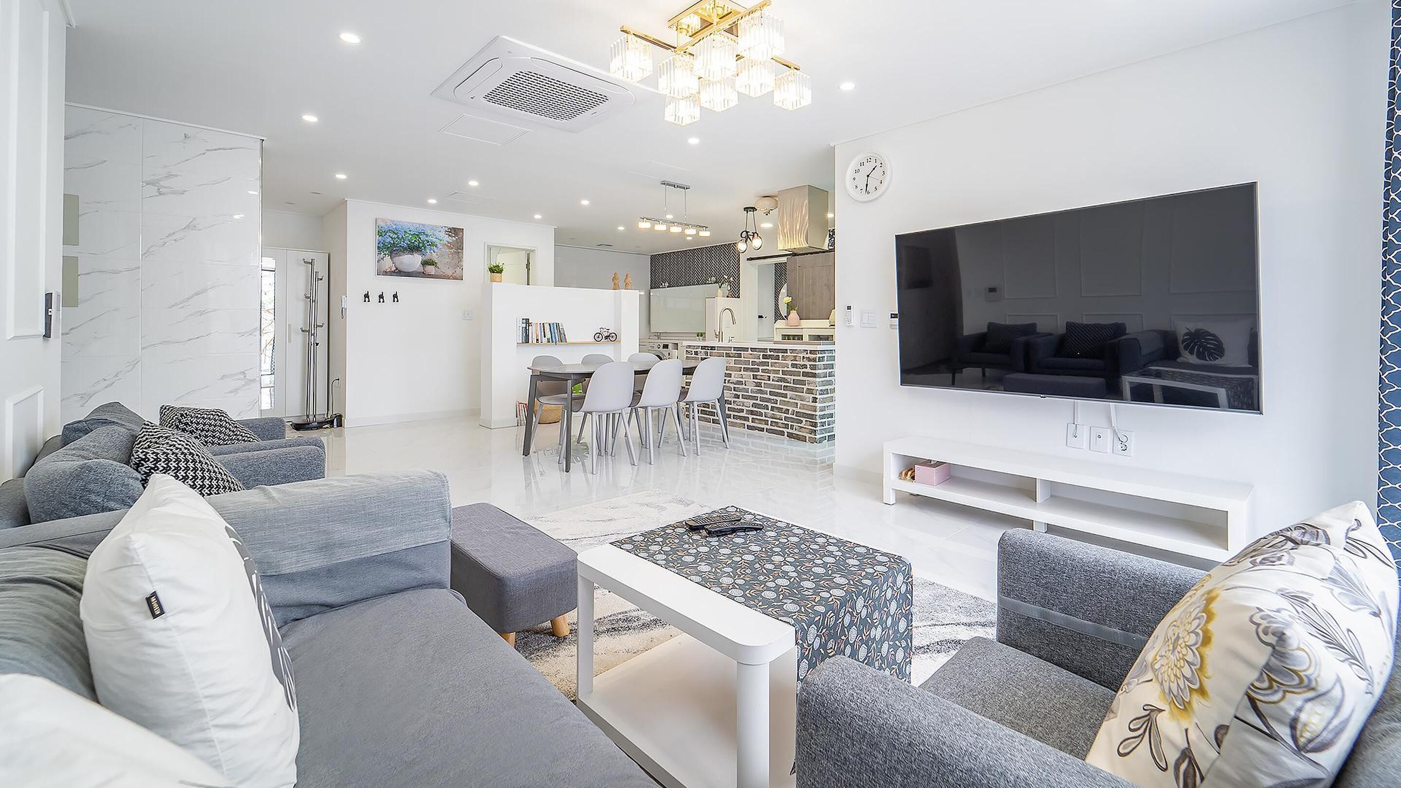 Una sala moderna con un sofá gris, un televisor de pantalla plana grande y un comedor con una cocina al fondo.