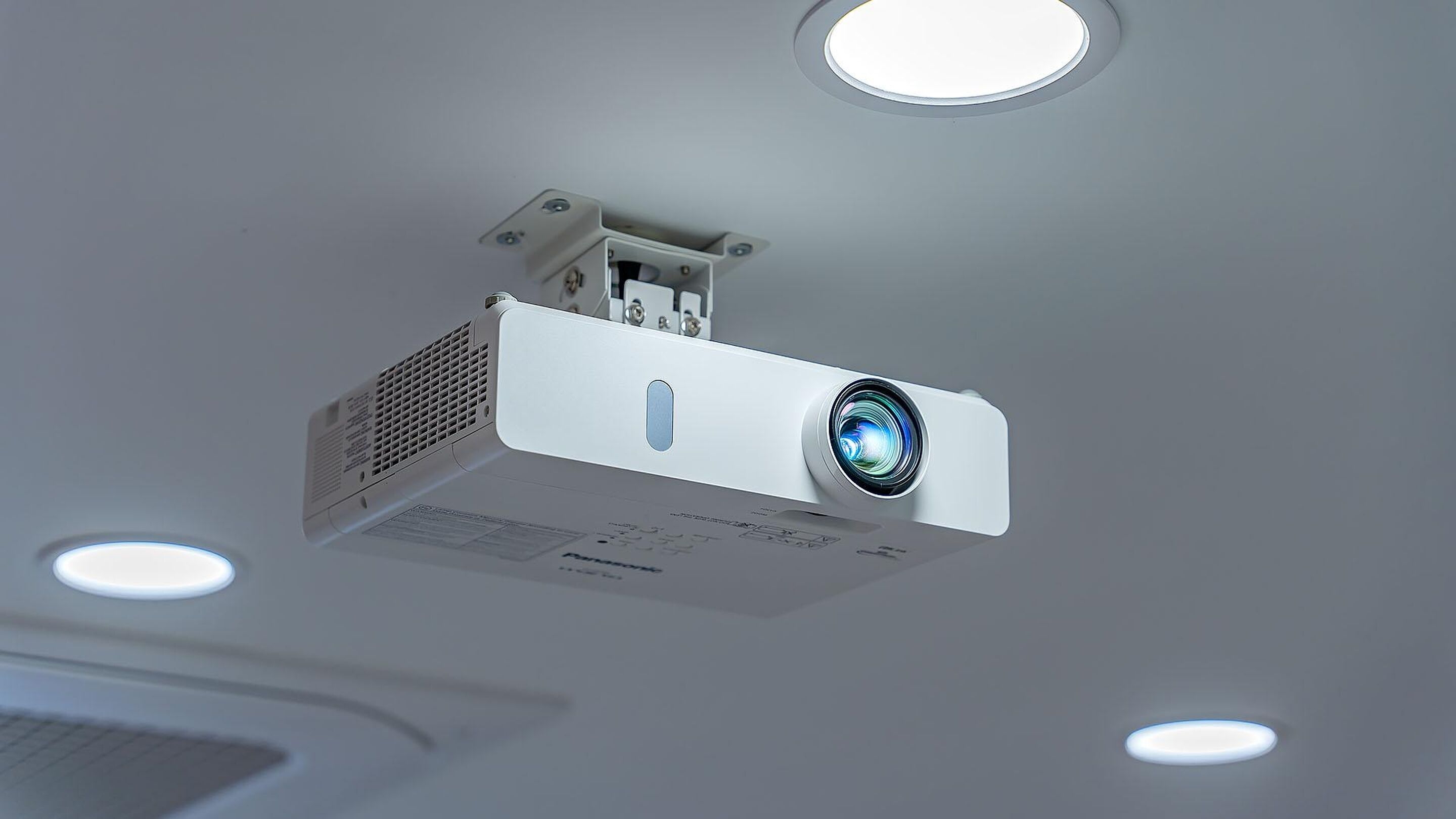 A ceiling-mounted projector with a lens and ventilation grille, mounted on a ceiling with recessed lighting.