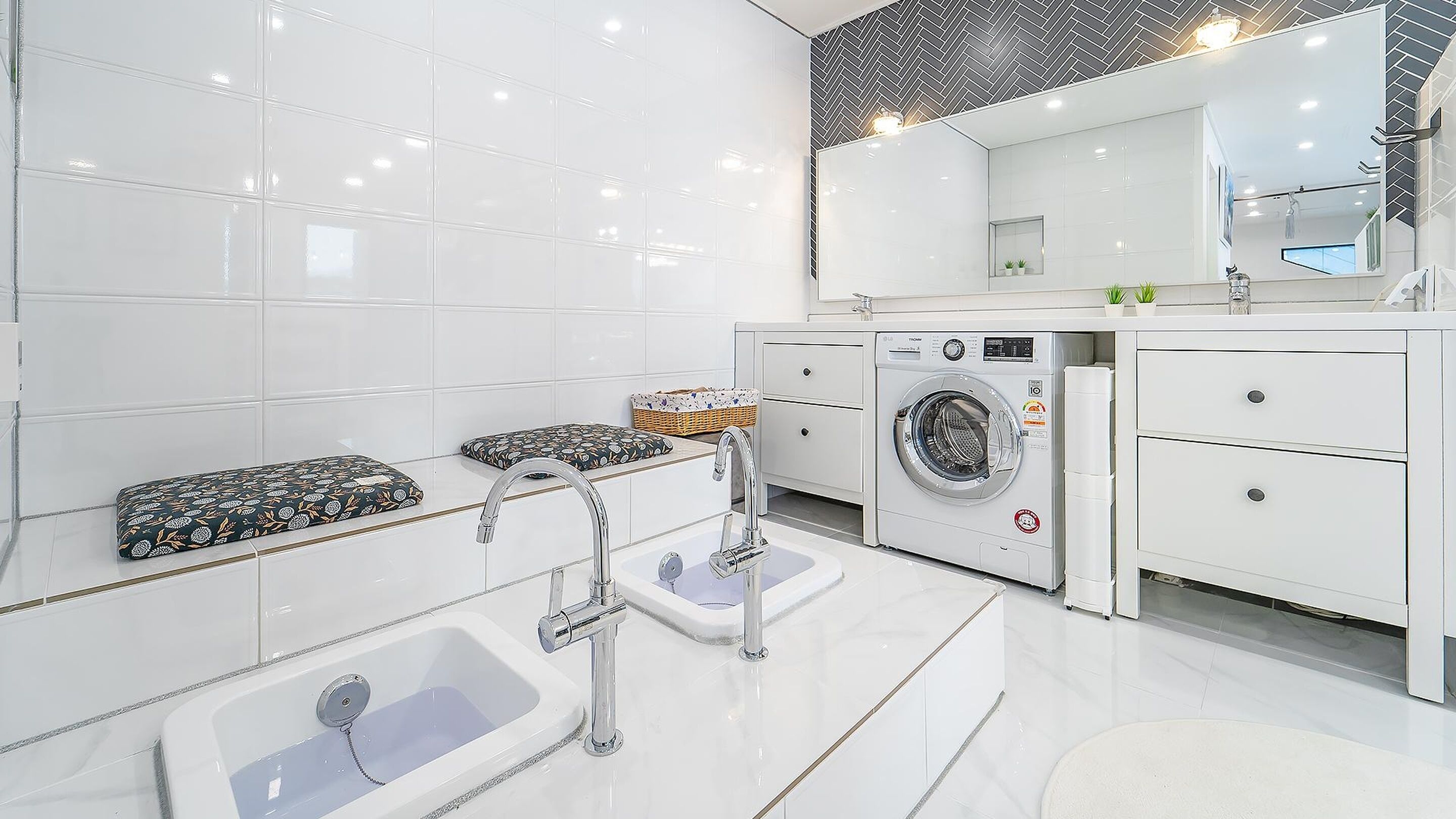 A modern bathroom with a large mirror, a washing machine, a bathtub, and two patterned poufs.