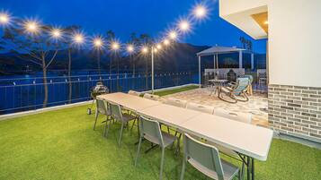 An outdoor patio with a long table and chairs, a grill, and string lights.