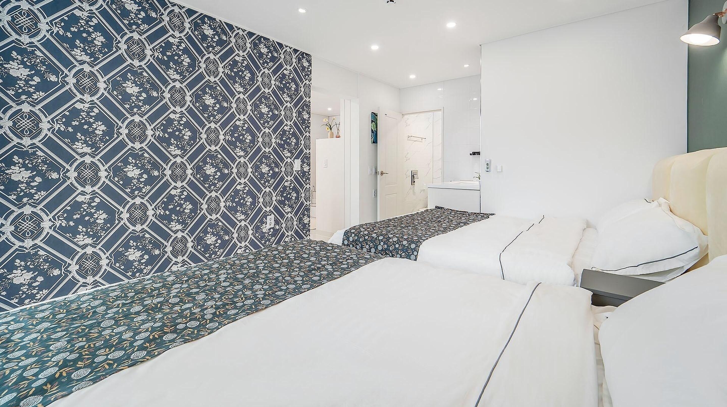 Ein modernes Hotelzimmer mit strukturiertem Wandpapier, einem Bett mit weißer Bettwäsche, einer kleinen Sitzecke und einem Badezimmer mit einer Glasdusche.