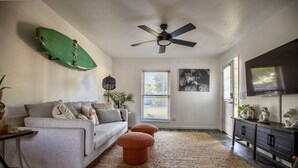 A living room with a sofa, ottomans, a ceiling fan, a TV, and artwork on the walls.