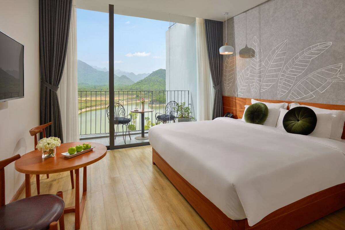 Quarto de hotel com uma cama grande, uma mesa pequena com um vaso de flores, uma cadeira e vista para um rio e montanhas através de uma varanda.