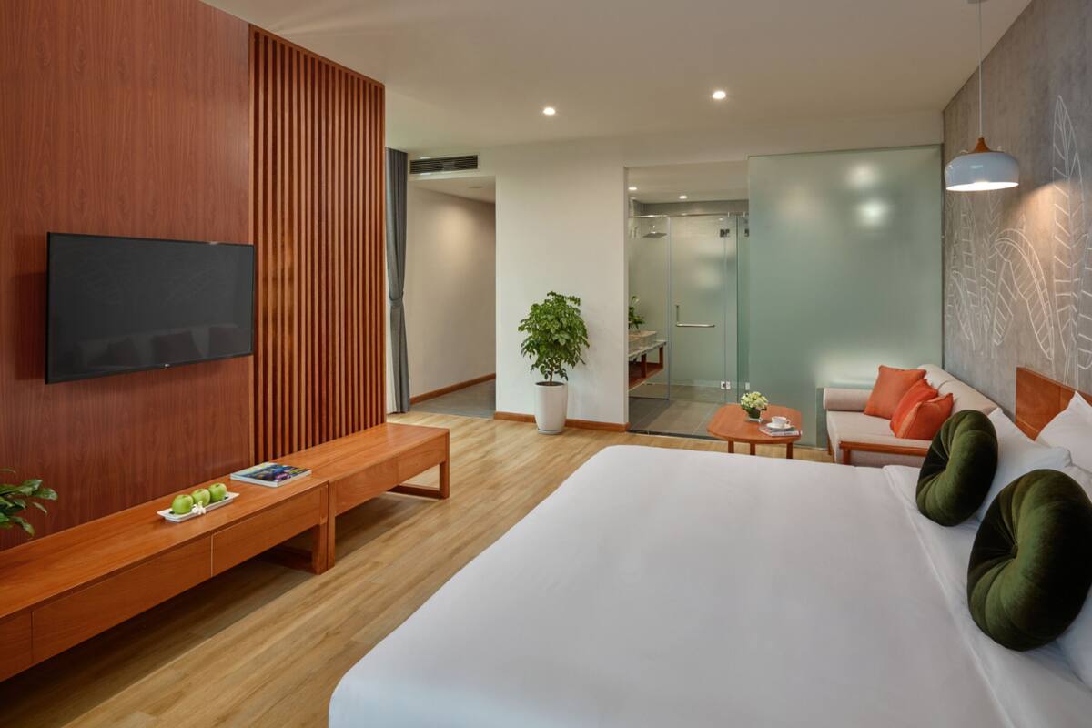 Quarto de hotel moderno com uma cama grande, um banco de madeira, uma televisão e uma área de estar com sofá e mesa.