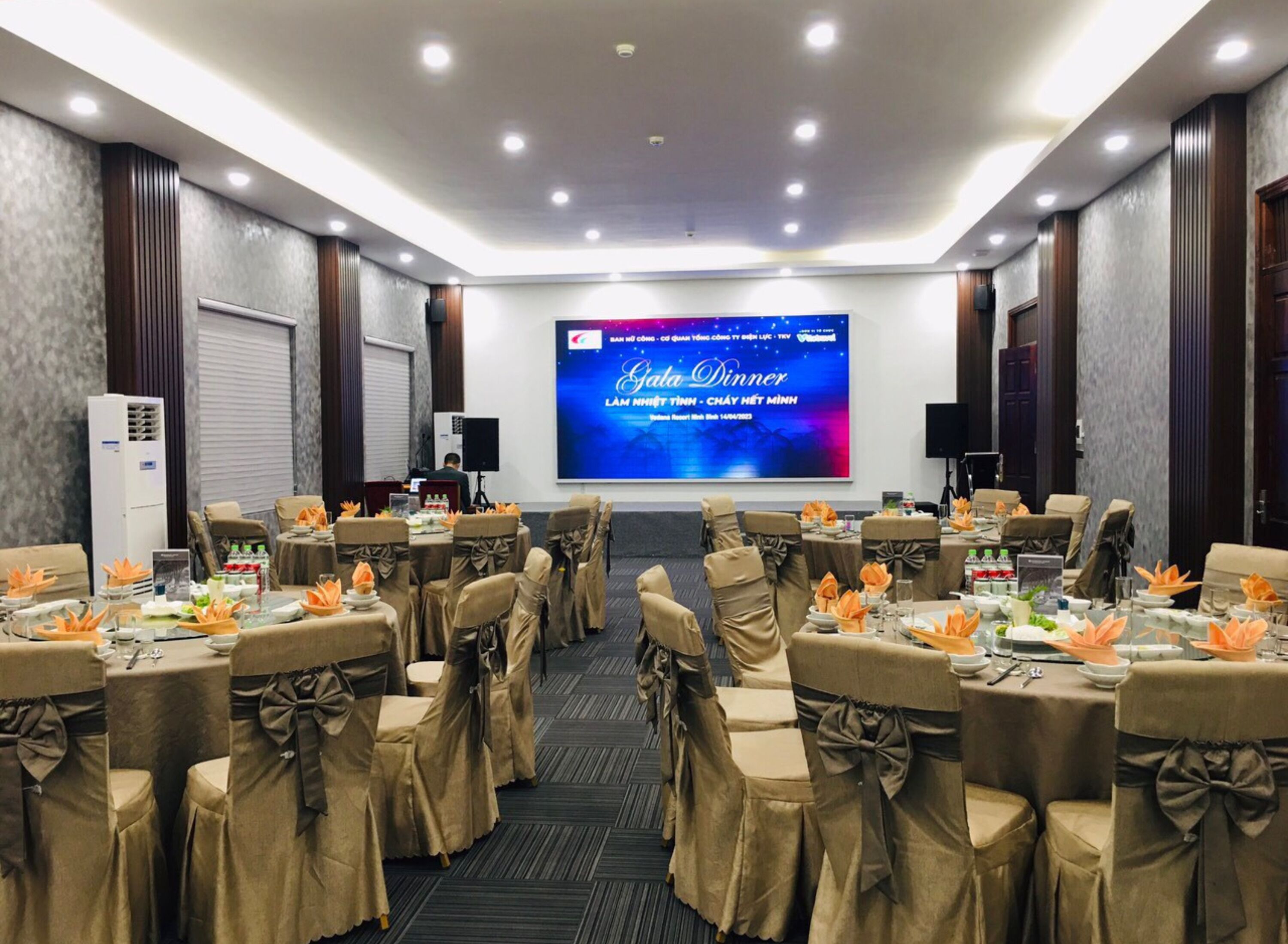 A conference room with tables set for a gala dinner, featuring a large screen displaying event details.