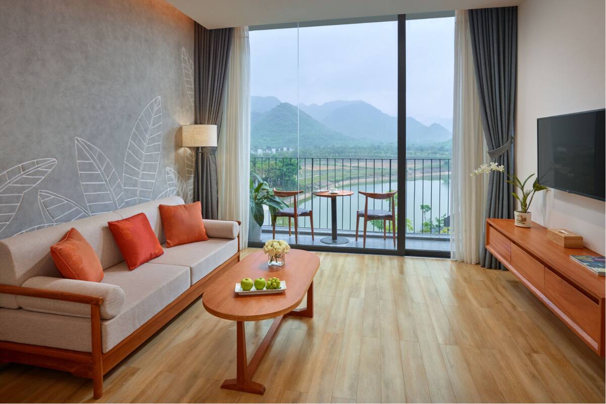 Quarto de hotel moderno com sofá, mesa de centro e varanda com vista para montanhas e um lago.