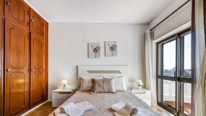 Une chambre avec un grand lit, une armoire en bois, deux tables de chevet avec des lampes, une fenêtre donnant sur un beau paysage et deux tableaux accrochés au mur.