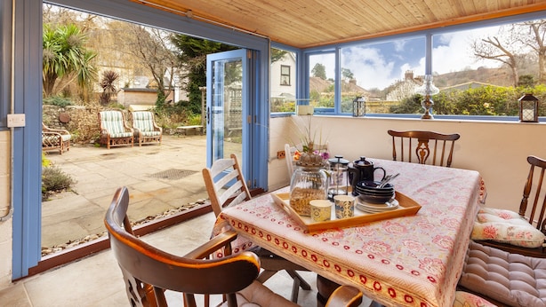 Un coin repas avec une table dressée pour deux personnes, des chaises et une vue sur le jardin grâce à de grandes fenêtres.