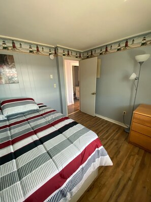 Un dormitorio con colcha a rayas, un aparador de madera, una lámpara y una puerta que da a otra habitación.