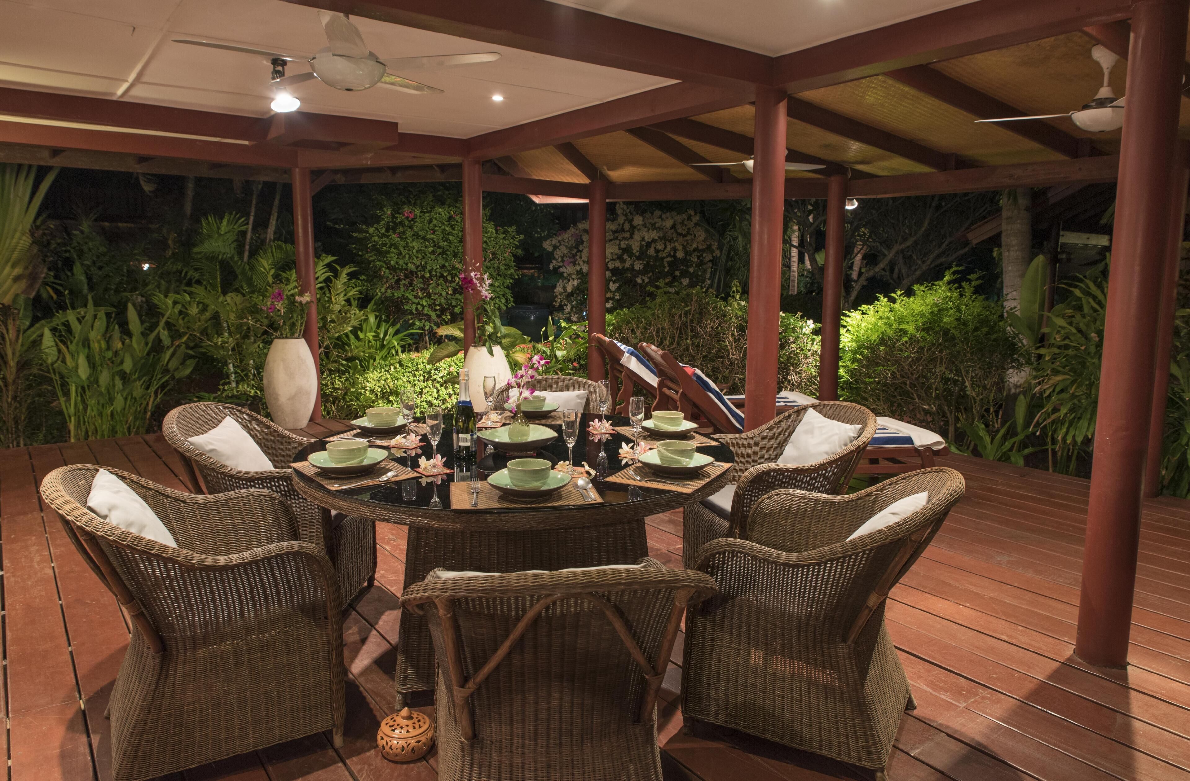 An outdoor dining area with wicker chairs and a round table set for four, surrounded by lush greenery and a wooden deck.