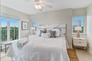 A bedroom with a large bed, a ceiling fan, two bedside tables with lamps, and a view of greenery through the windows. - Bright, Airy Oceanview Condo on Amelia Island! (Amelia Island)