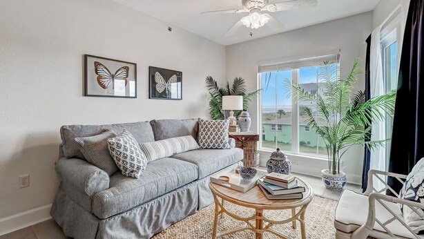 A living room with a grey sofa, two butterfly wall art pieces, a ceiling fan, and a large window with a view of buildings. - Bright, Airy Oceanview Condo on Amelia Island! (Amelia Island)